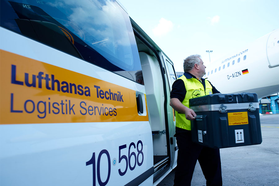 Ein Lufthansa-Mitarbeiter in einer Lufthansa Warnweste trägt eine Box aus einem Lieferwagen zu einem Lufthansa-Flugzeug, um sie umzuladen