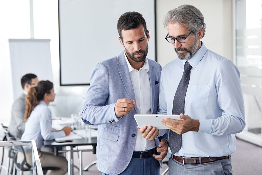 Bild einer Bürosituation, in der zwei Männer im Anzug eine Besprechung mit einem Tablet abhalten, was die moderne Digitalisierung im Fuhrparkmanagement verdeutlicht. Die Nutzung von Technologie ermöglicht eine effizientere Verwaltung und erleichtert die Konzentration auf strategische Flottenmanagementaufgaben.